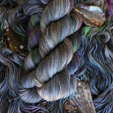 DK yarn colored with different shades of grey and streaks of green, and dark pink, also there are also blue and purple speckles. These skeins are on top of unwound skeins of the same color way with some shells on top.