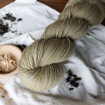 A very calm pretty light brownish green colored DK yarn skein on top of a white tablecloth and rice grains and soil in the corner on top of a wooden table.
