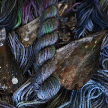 DK mini yarn colored with different shades of grey and streaks of green, and dark pink, also there are also blue and purple speckles. These skeins are on top of unwound skeins of the same color way with some shells on top.