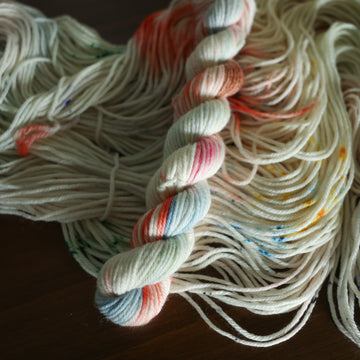 DK mini white yarn with mixed colors of light pink purple and red/orange and brown with speckles, on top of an unwound version of that same yarn. all on top of a dark wooden table.