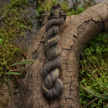 A dark grey DK mini yarn with hints of green colors, on top of a tree limb that is hovering over a pond, with different plants around the pond, and around the yarn.