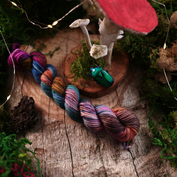 DK-mini yarn colored with a hot pink, light blue, dark blue/purple, with some green and orange/brown colors, on a wooden table with some moss surrounding it and a wooden circle with mushrooms on it and a small glass beetle.