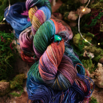 DK yarn colored with a hot pink, light blue, dark blue/purple, with some green and orange/brown colors, on top of the same yarn but unwound, with moss underneath and some fairy lights around the moss and a small glass looking beetle.