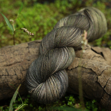 A dark grey DK yarn with hints of green colors, on top of a tree limb that is hovering over a pond, with different plants around the pond, and around the yarn.