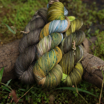 Different DK yarn skeins, one colored a lime green and grey, one colored a dark grey, and one colored green with blue speckles, on top of a tree limb that is hovering over a pond, with different mossy plants around the pond, and around the yarn.