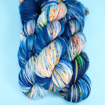 DK yarn colored with different shades of blue and white with multicolored speckles on the portion of the yarn that Is white. all on top of a light blue background.