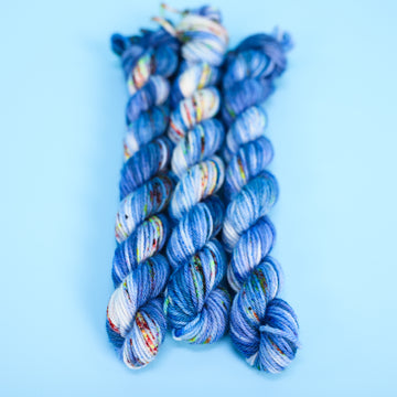 DK mini yarn colored with different shades of blue and white with multicolored speckles on the portion of the yarn that Is white. all on top of a light blue background.