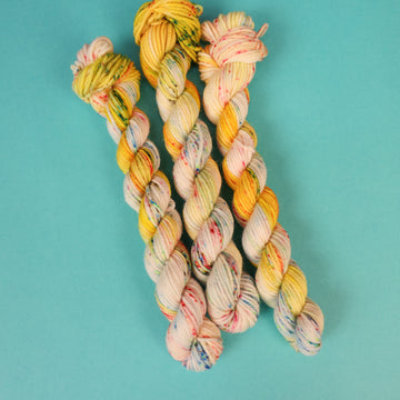 A light cream colored DK mini yarn with different colored speckles all over and banana colored yellow at the top of the skein on top of a light blue background.