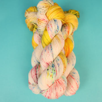 A light cream colored DK yarn with different colored speckles all over and banana colored yellow at the top of the skein on top of a light blue background.