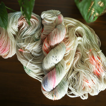 DK white yarn with mixed colors of light pink purple and red/orange and brown with speckles, on top of an unwound version of that same yarn. all on top of a dark wooden table with a plant overhead in a gold pot.