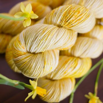 A yellow varied Sparkle Sock skein sitting on top of three yellow varied DK skeins, all sitting on top of a brown table surrounded by yellow flowers.
