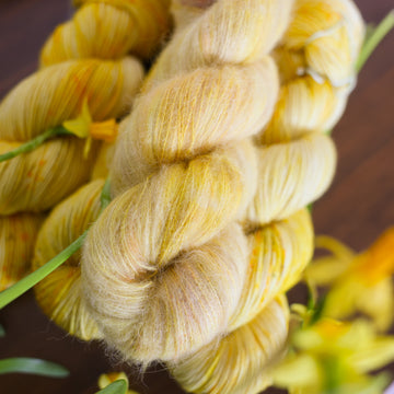 A yellow varied Suri skein sitting on top 3 yellow varied DK skeins all sitting on top of a brown table surrounded by yellow flowers.