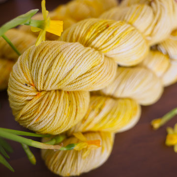 A yellow varied Worsted skein sitting on top 3 yellow varied DK skeins, all sitting on top of a brown table surrounded by yellow flowers.
