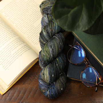 A dark blue, green, and black speckled yarn next to an open book, two closed books with glasses on top, and a plant overhead of the yarn.