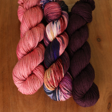 Darkness Lovely Kit sitting on top of a table. From left to right the skeins are light pink, a multicolored skein with purple red and blue streaks, and deep purple.