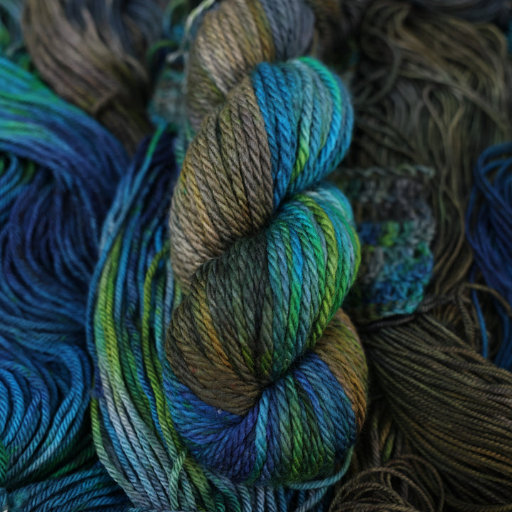 Bulky yarn colored with dark brown dark blue and green next to crochet and knit swatches on this yarn. This is all on top of unwound skeins of this same color way.