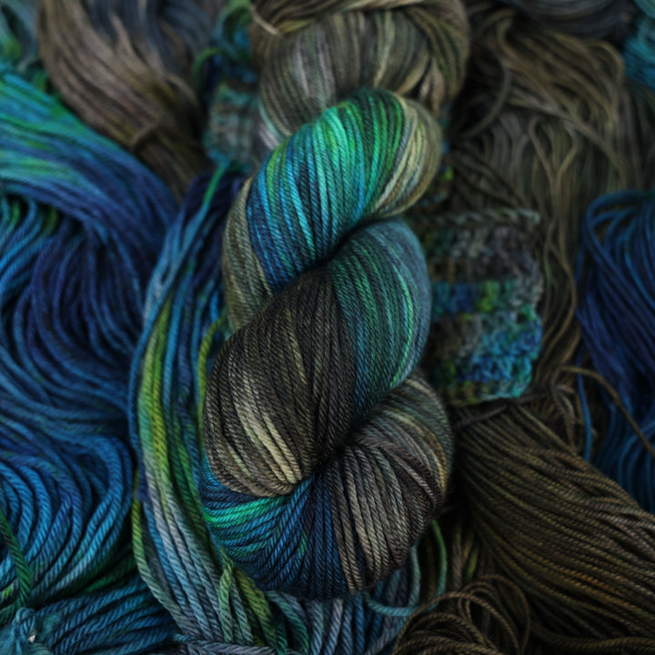 DK yarn colored with dark brown dark blue and green next to crochet and knit swatches on this yarn. This is all on top of unwound skeins of this same color way.