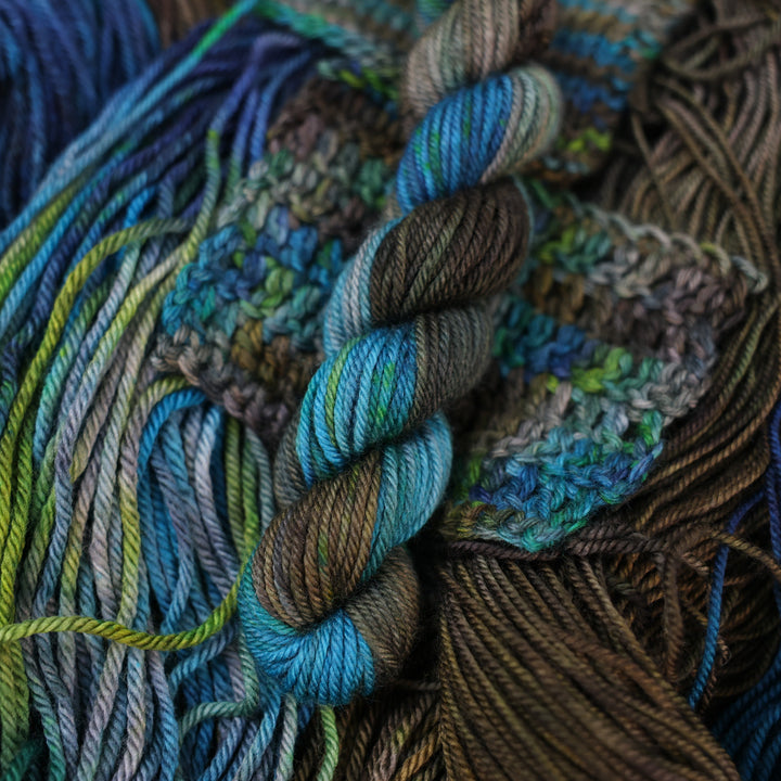 DK mini yarn colored with dark brown dark blue and green next to crochet and knit swatches on this yarn. This is all on top of unwound skeins of this same color way.