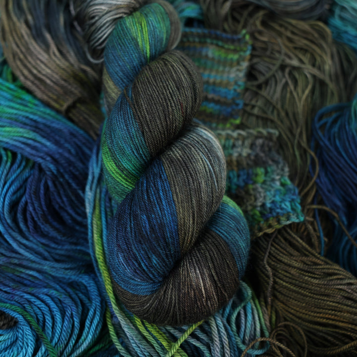 Fingering yarn colored with dark brown dark blue and green next to crochet and knit swatches on this yarn. This is all on top of unwound skeins of this same color way.