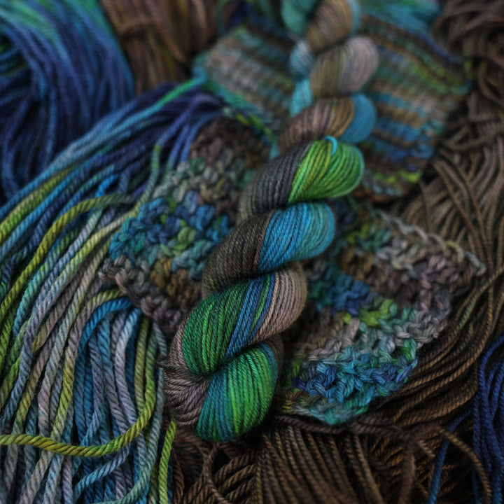 Fingering mini yarn colored with dark brown dark blue and green next to crochet and knit swatches on this yarn. This is all on top of unwound skeins of this same color way.