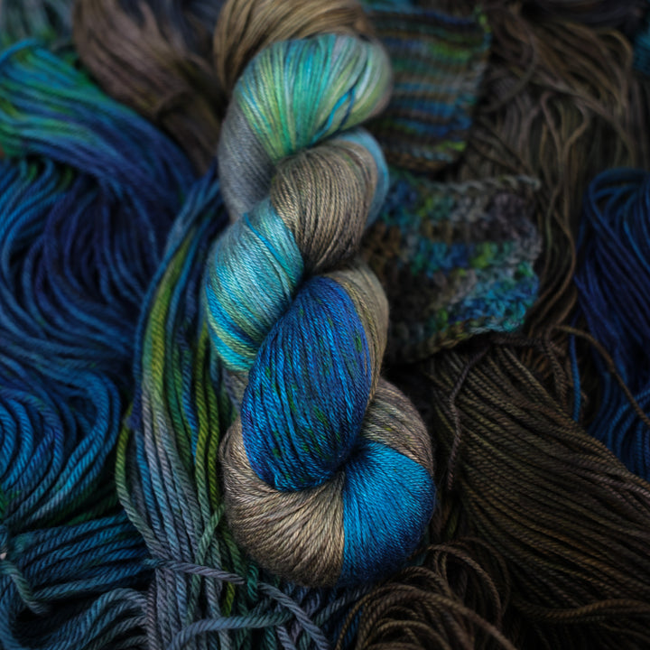 Silk yarn colored with dark brown dark blue and green next to crochet and knit swatches on this yarn. This is all on top of unwound skeins of this same color way.