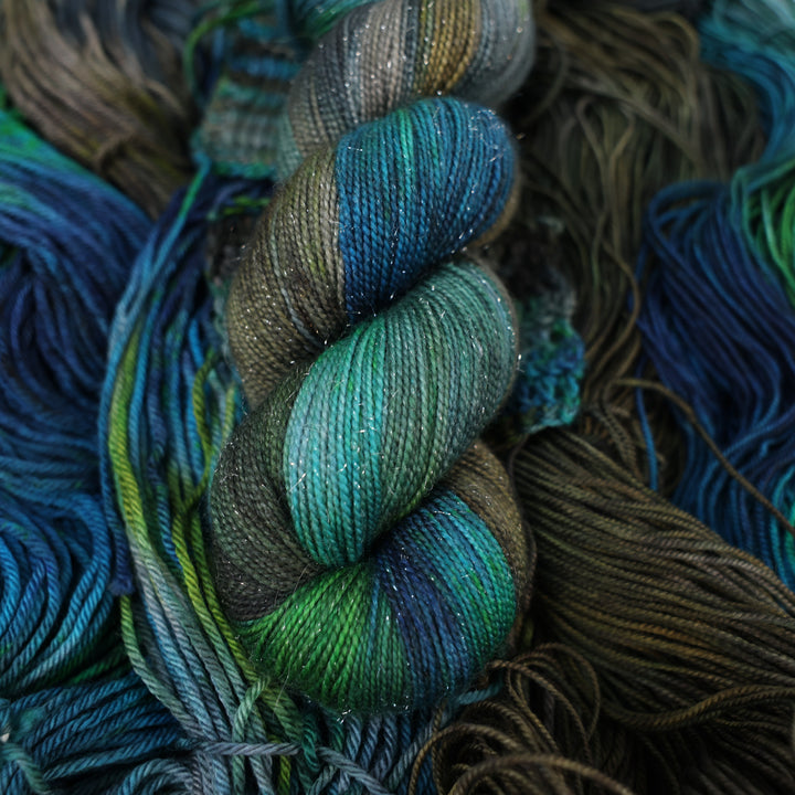 Sparkle DK yarn colored with dark brown dark blue and green next to crochet and knit swatches on this yarn. This is all on top of unwound skeins of this same color way.
