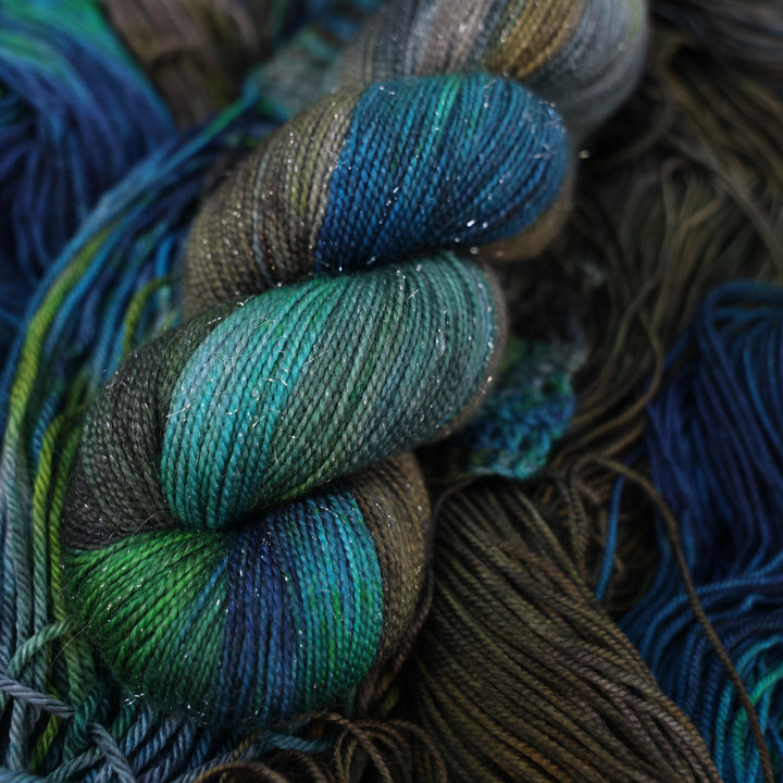Sparkle fingering yarn colored with dark brown dark blue and green next to crochet and knit swatches on this yarn. This is all on top of unwound skeins of this same color way.