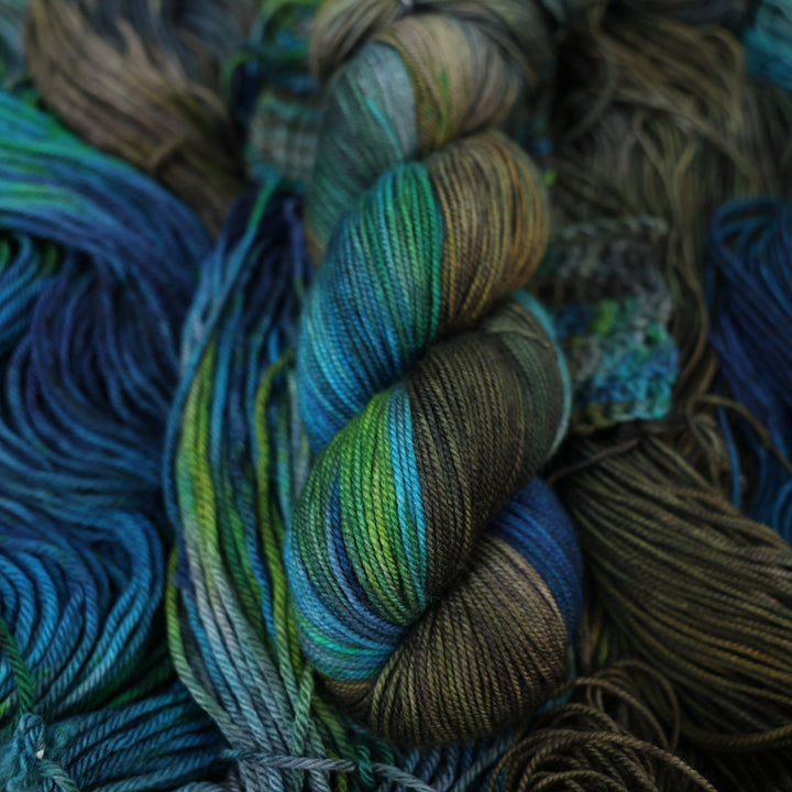 Sport yarn colored with dark brown dark blue and green next to crochet and knit swatches on this yarn. This is all on top of unwound skeins of this same color way.