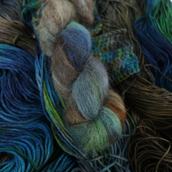 Suri yarn colored with dark brown dark blue and green next to crochet and knit swatches on this yarn. This is all on top of unwound skeins of this same color way.