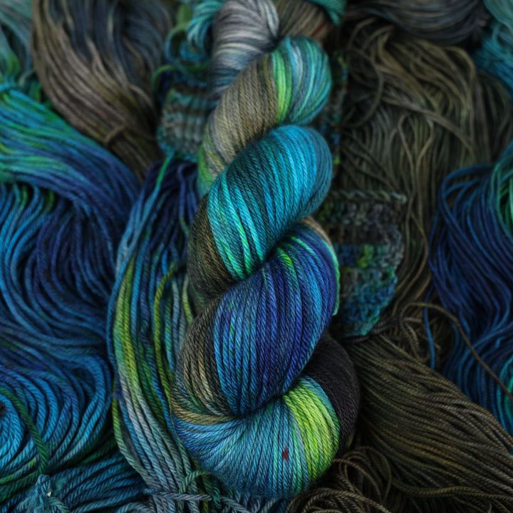 Worsted yarn colored with dark brown dark blue and green next to crochet and knit swatches on this yarn. This is all on top of unwound skeins of this same color way.
