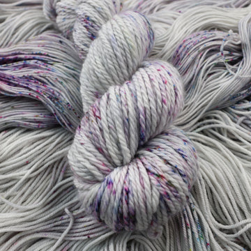 Bulky yarn colored a light grey with some pink, purple, and blue speckles. This skein is on top of unwound yarn of the same color way.
