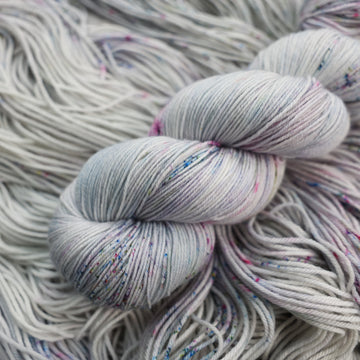Fingering yarn colored a light grey with some pink, purple, and blue speckles. This skein is on top of unwound yarn of the same color way.
