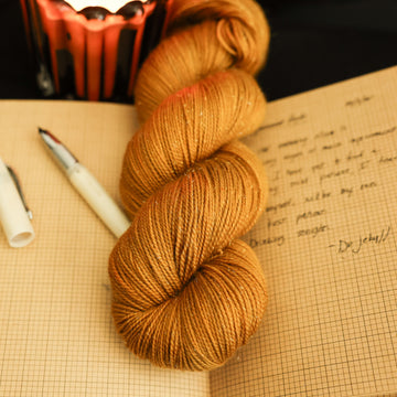 A light burnt orange sparkle yarn on top of an open notebook that has a letter written on it with an open ink pen next to it and a lit orange candle in the back, all on a black background