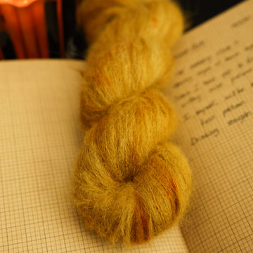 A light burnt orange suri yarn on top of an open notebook that has a letter written on it and a lit orange candle in the back, all on a black background