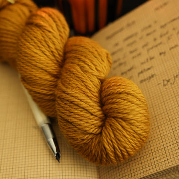 A light burnt orange bulky yarn on top of an open notebook with an ink pen next to it and a lit orange candle in the back, all on a black background