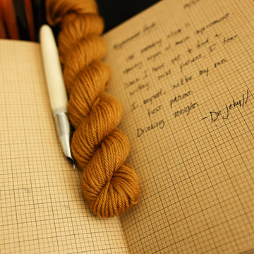 A light burnt orange dk mini yarn on top of an open notebook that has a letter written on it with an ink pen next to it and a lit orange candle in the back, all on a black background