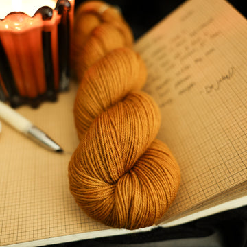 A light burnt orange fingering yarn on top of an open notebook that has a letter written on it with an ink pen next to it and a lit orange candle on the book, all on a black background