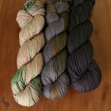 Edgar Allen Poe Kit sitting on top of a wooden table. Skeins from left to right are white with green speckles, grey with green and brown streaks, and dark grey with dark green streaks.