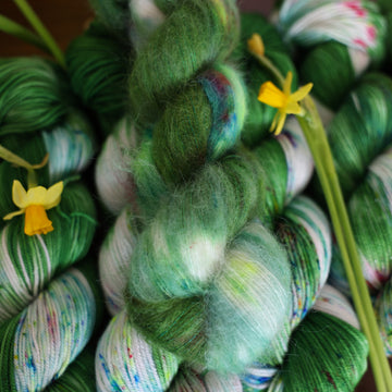 Wound up Alpaca yarn that is colored with a darkish green with some lighter shades mixed in, and a white streak coming through the green on both sides that has different colored speckles with light greens pinks and blues. There are yellow flowers on top of the yarn.