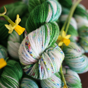 Wound up Bulky yarn that is colored with a darkish green with some lighter shades mixed in, and a white streak coming through the green on both sides that has different colored speckles with light greens pinks and blues. There are yellow flowers on top of the yarn.