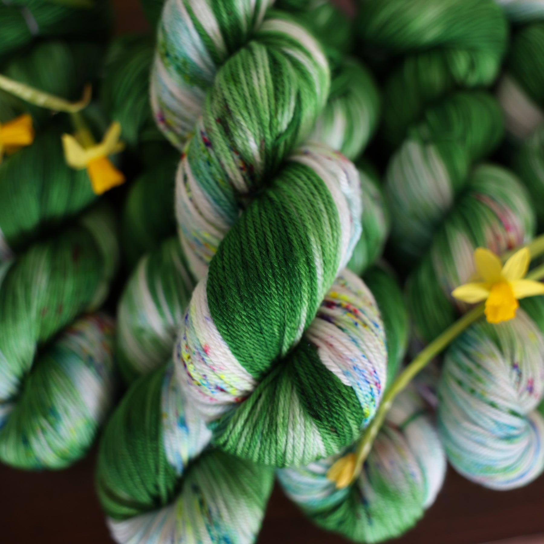 Wound up DK yarn that is colored with a darkish green with some lighter shades mixed in, and a white streak coming through the green on both sides that has different colored speckles with light greens pinks and blues. There are yellow flowers on top of the yarn.