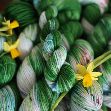Wound up DK mimi yarn that is colored with a darkish green with some lighter shades mixed in, and a white streak coming through the green on both sides that has different colored speckles with light greens pinks and blues. There are yellow flowers on top of the yarn.