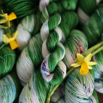 Wound up Fingering mini yarn that is colored with a darkish green with some lighter shades mixed in, and a white streak coming through the green on both sides that has different colored speckles with light greens pinks and blues. There are yellow flowers on top of the yarn.