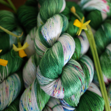 Wound up Silk yarn that is colored with a darkish green with some lighter shades mixed in, and a white streak coming through the green on both sides that has different colored speckles with light greens pinks and blues. There are yellow flowers on top of the yarn.