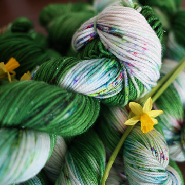 Wound up Sparkle DK yarn that is colored with a darkish green with some lighter shades mixed in, and a white streak coming through the green on both sides that has different colored speckles with light greens pinks and blues. There are yellow flowers on top of the yarn.