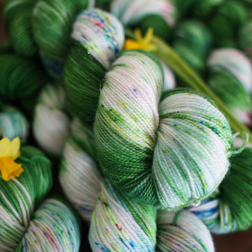 Wound up Sock yarn that is colored with a darkish green with some lighter shades mixed in, and a white streak coming through the green on both sides that has different colored speckles with light greens pinks and blues. There are yellow flowers on top of the yarn.