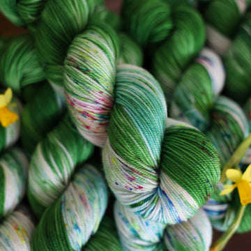 Wound up Sport yarn that is colored with a darkish green with some lighter shades mixed in, and a white streak coming through the green on both sides that has different colored speckles with light greens pinks and blues. There are yellow flowers on top of the yarn.