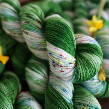 Wound up Worsted yarn that is colored with a darkish green with some lighter shades mixed in, and a white streak coming through the green on both sides that has different colored speckles with light greens pinks and blues. There are yellow flowers on top of the yarn.