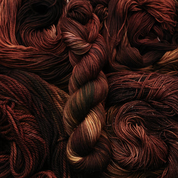 A dark brown colored Sport yarn that has some lighter tones of brown and different shades. There are unwound versions of this skein in different weights underneath this skein.