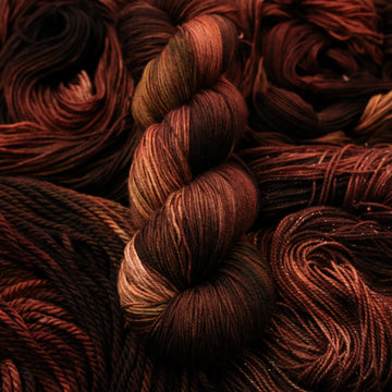 A dark brown colored Fingering yarn that has some lighter tones of brown and different shades. There are unwound versions of this skein in different weights underneath this skein.